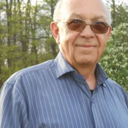 Portret starszego mężczyzny w okularach przeciwsłonecznych, ubranego w koszulę w paski, na tle rozkwitającego drzewa i zieleni.