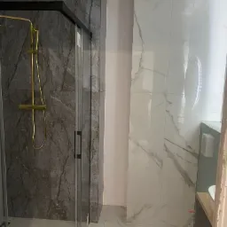 Kabina prysznicowa z czarną ramą i złotą armaturą na tle ściany wyłożonej płytkami imitującymi ciemny marmur, obok ściana z białych płytek marmurowych z okrągłym lustrem i wentylatorem.