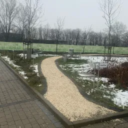 Kamyczkowa ścieżka wijąca się przez park z ławkami i młodymi drzewami, w tle zielony płot z logo West Forum, częściowo pokryta śniegiem, widoczne tabliczki z nazwami firm.