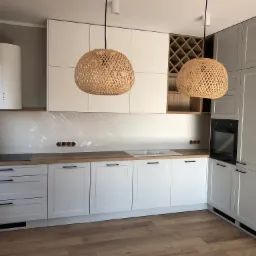 Jasna kuchnia w stylu skandynawskim z białymi szafkami z frezowanymi frontami, drewnianym blatem i dwoma plecionymi lampami wiszącymi, widok na narożną zabudowę i ekspozycję na wino.