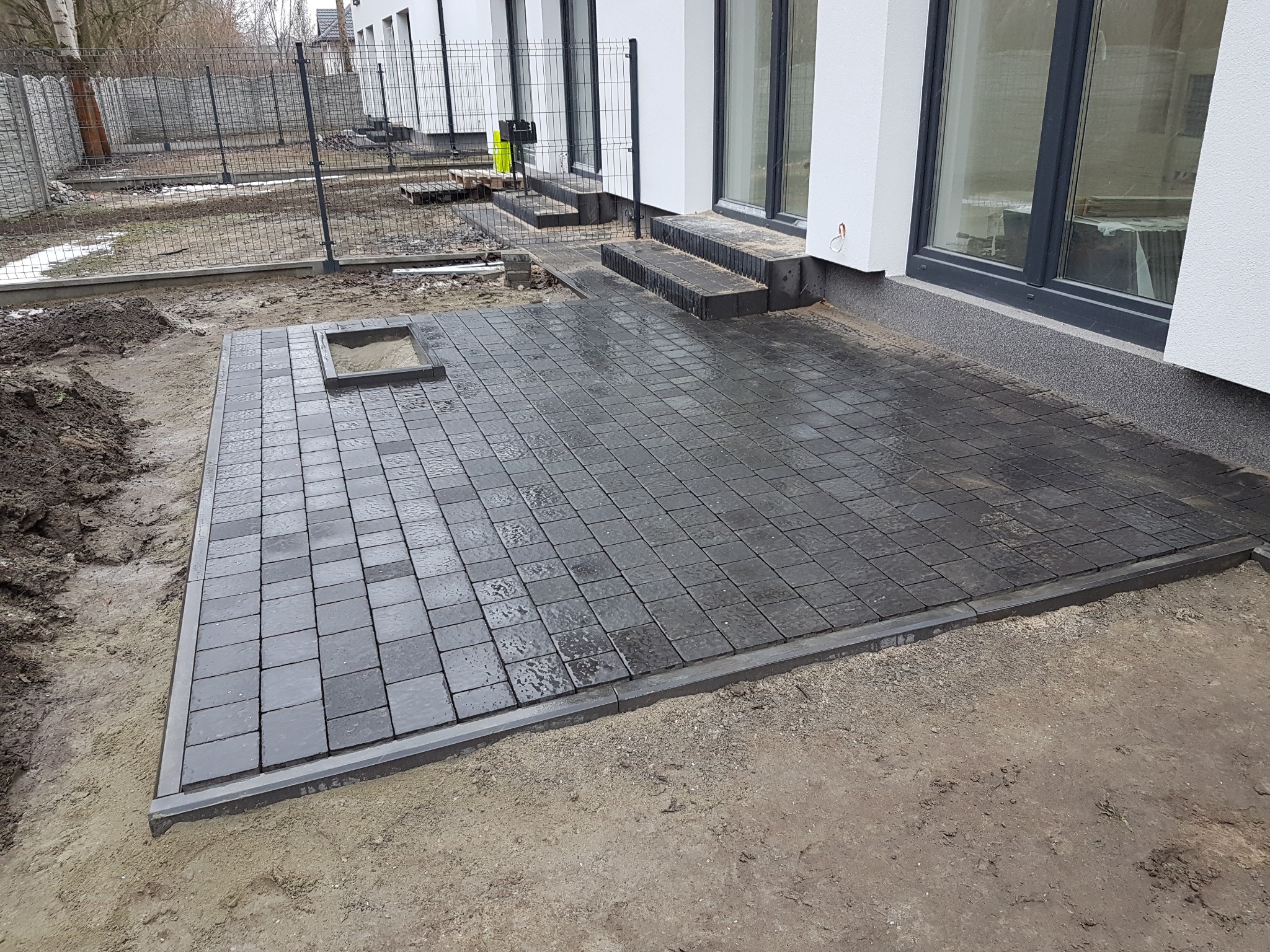 Nowo ułożony, ciemnoszary taras z kostki brukowej na osiedlu, z widocznym otworem rewizyjnym i schodami prowadzącymi do budynku.
