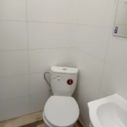 Biała toaleta i umywalka w małej łazience wyłożonej białymi, kwadratowymi płytkami, widoczne także fragment lustra i drewnopodobnej podłogi.