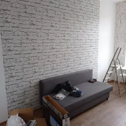 Pokój po odświeżeniu z tapetą imitującą białą cegłę na ścianie, szara sofa z narzędziami, drabina oparta o ścianę, kartony na podłodze, widok na fragment pokoju po remoncie.