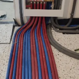 Rozdzielacz ogrzewania podłogowego z rurami w kolorze czerwonym i niebieskim, wychodzącymi z białej szafki, widoczne fragmenty podłogi w trakcie montażu.