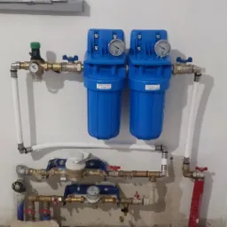 Dwa niebieskie filtry wody z manometrami zamontowane na ścianie w Chełmie, połączone rurami z mosiądzu i białego plastiku, widoczne również wodomierze i zawory.