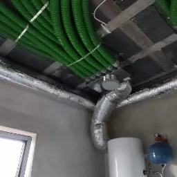 Widok z dołu na montaż instalacji wentylacyjnych z zielonymi rurami karbowanymi pod sufitem oraz aluminiowymi przewodami wentylacyjnymi, widoczny bojler i naczynie wzbiorcze w surowym wnętrzu.