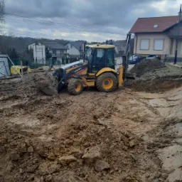 Żółta koparko-ładowarka Volvo BL71B na placu budowy, widoczne świeżo wykopane rowy w ziemi, w tle domy mieszkalne i pochmurne niebo.
