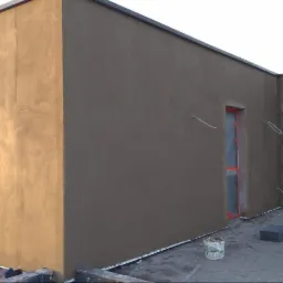 Ocieplona elewacja budynku z widocznymi oknami zabezpieczonymi folią, świeżo nałożony tynk w kolorze beżowym i szarym, widoczne ślady po szpachlowaniu.