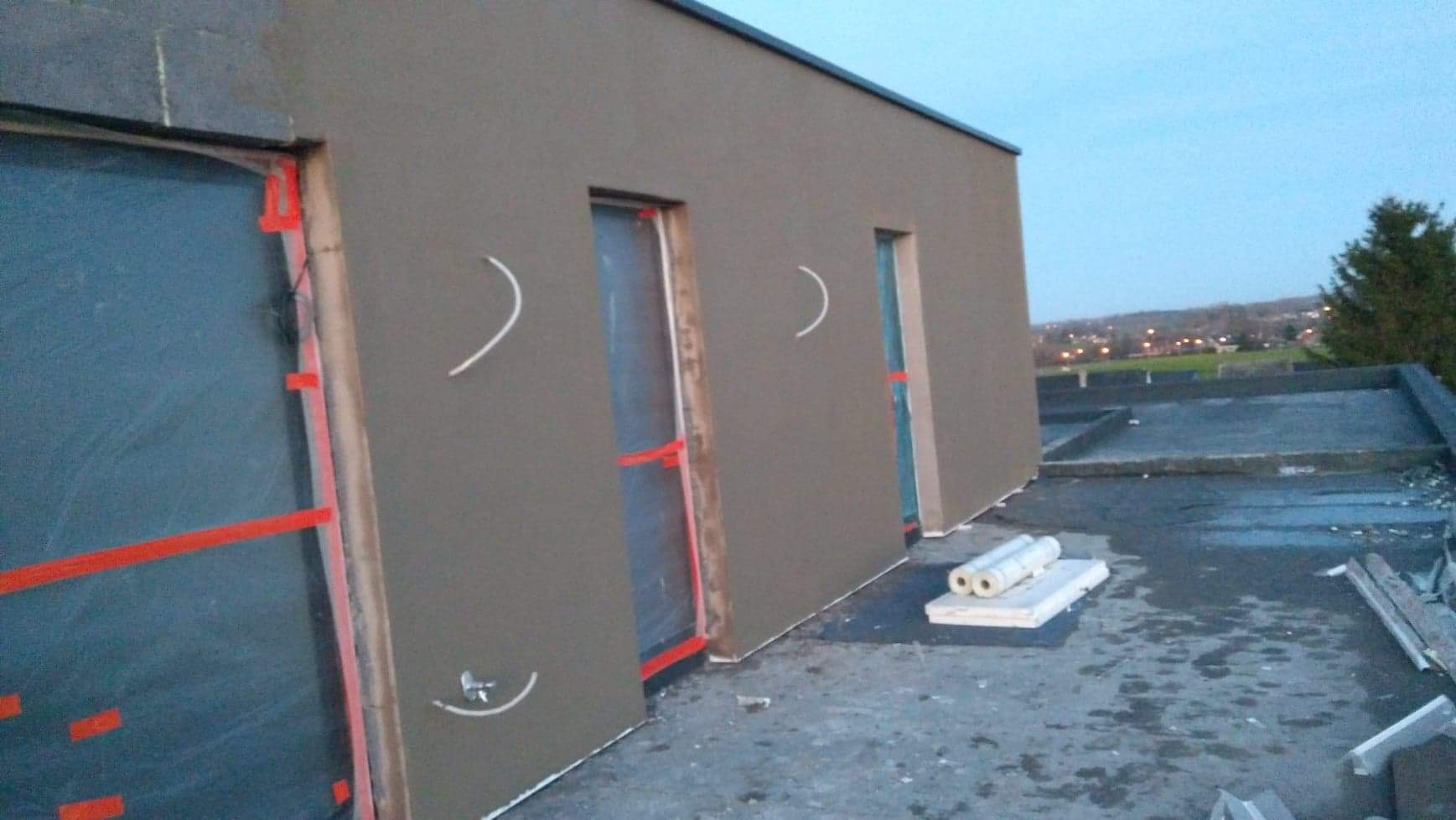 Budynek w trakcie ocieplania elewacji, widoczne okna zabezpieczone folią, świeżo nałożony tynk zewnętrzny w kolorze szarym, elementy rusztowania i rolki izolacji na dachu.