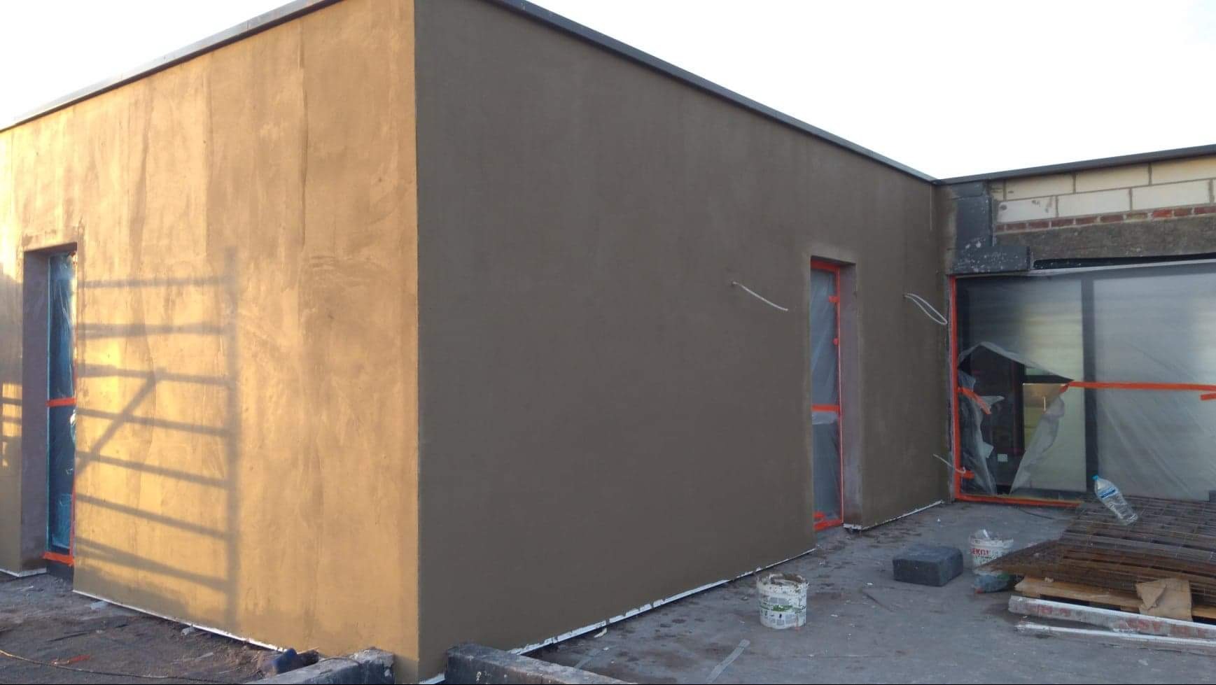 Ocieplona elewacja budynku z widocznymi oknami zabezpieczonymi folią, świeżo nałożony tynk w kolorze beżowym i szarym, widoczne ślady po szpachlowaniu.