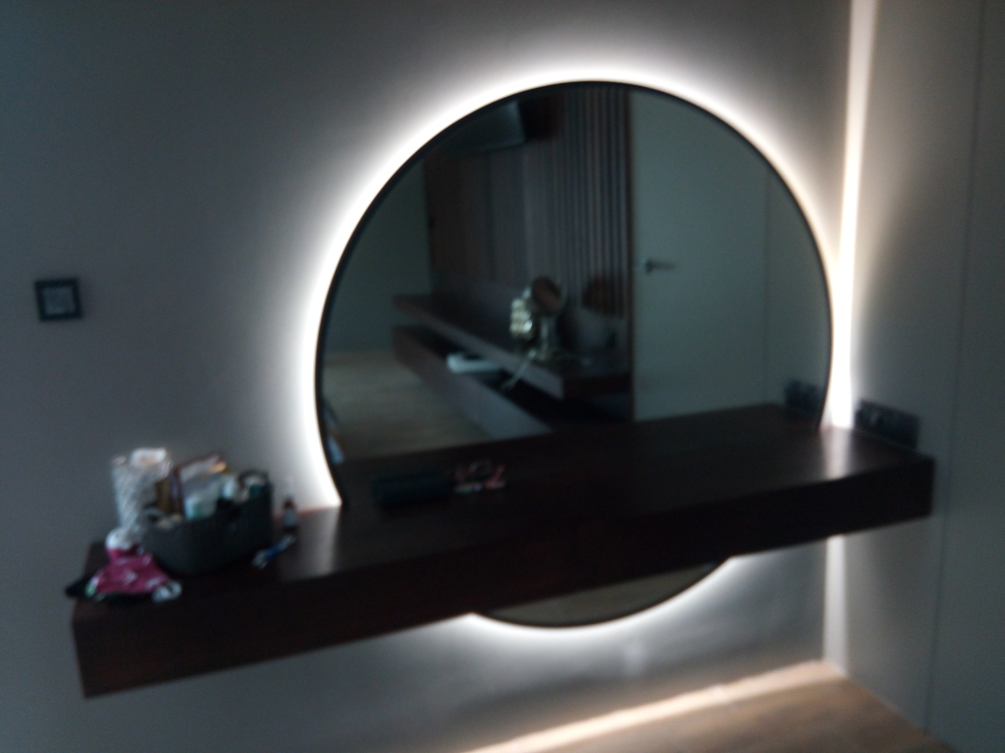 Okrągłe lustro z podświetleniem LED, zamontowane nad wiszącą, ciemną półką z drobnymi przedmiotami. W tle widoczne odbicie fragmentu wnętrza.