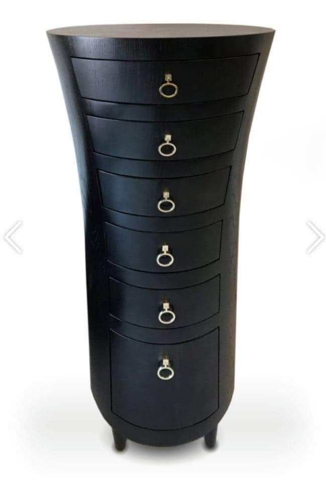 Wysoka, czarna komoda z sześcioma szufladami o nietypowym, zakrzywionym kształcie i złotych uchwytach, prezentowana w Warszawie.