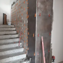 Układanie ciemnych, brązowo-szarych płytek na ścianie przy schodach, z użyciem poziomicy laserowej i klipsów poziomujących, widoczny częściowo pomalowany schodek.