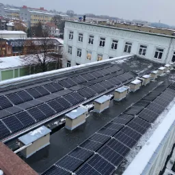 Instalacja paneli słonecznych na dachu budynku w Gliwicach, widoczne kominy wentylacyjne i pracownicy na dachu w tle, zimowa sceneria.