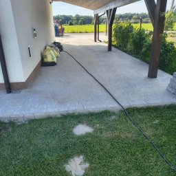 INSTAL-BRUK Sebastian Raba - Ułożona kostka brukowa na podjeździe z drewnianą wiatą. W tle zieleń i pracownicy. Widoczne worki z materiałem i kabel zasilający. Kąt ujęcia z poziomu trawnika.