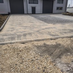 INSTAL-BRUK Sebastian Raba - Ułożona szara kostka brukowa przed budynkiem magazynowym z szarymi bramami. Część powierzchni wyłożona ażurowymi płytami. Na pierwszym planie żwir.