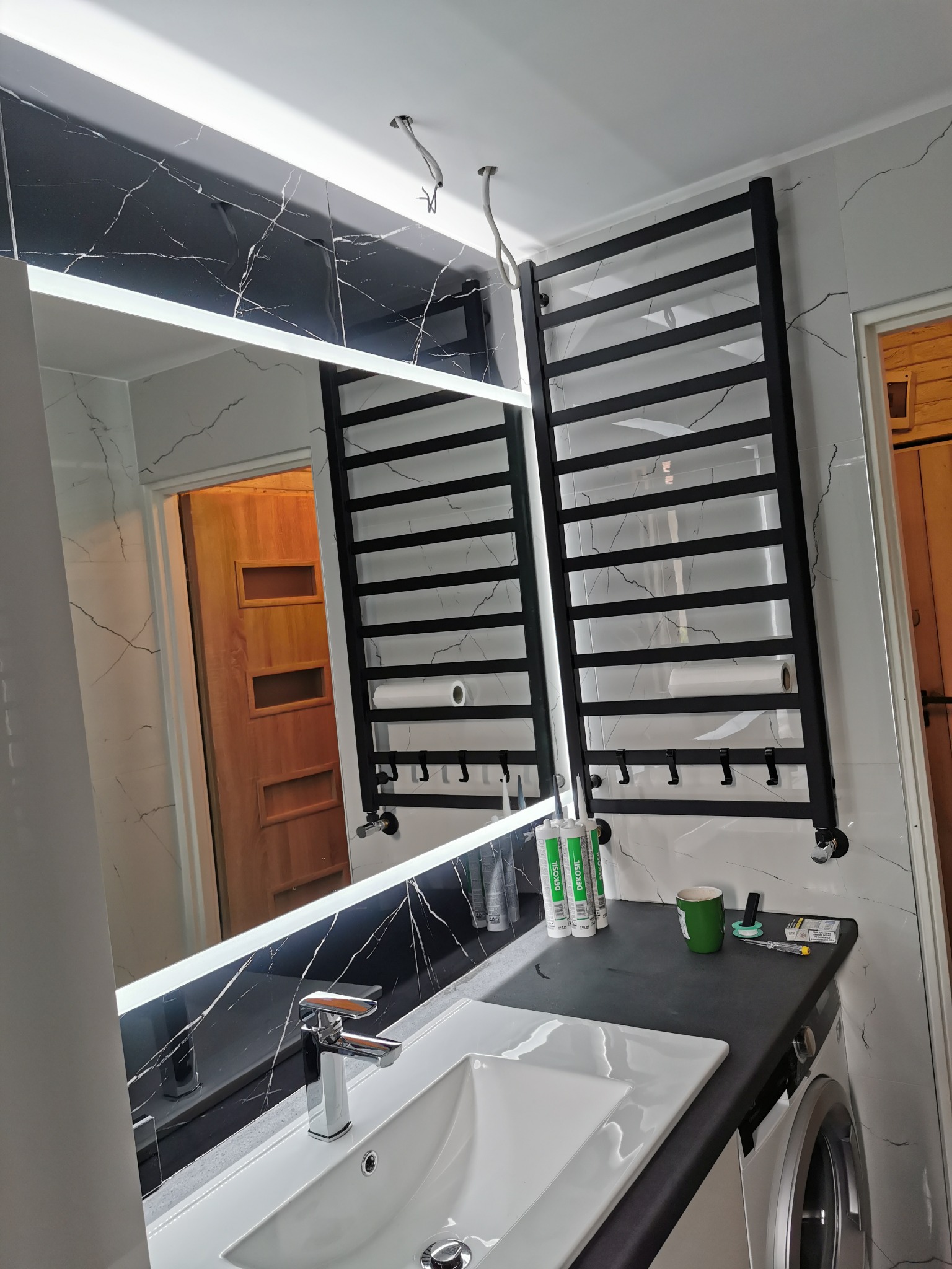 Nowoczesna łazienka po remoncie: biała umywalka z chromowaną baterią, podświetlane lustro na ścianie z czarnymi płytkami imitującymi marmur, czarny grzejnik drabinkowy, akcesoria montażowe...