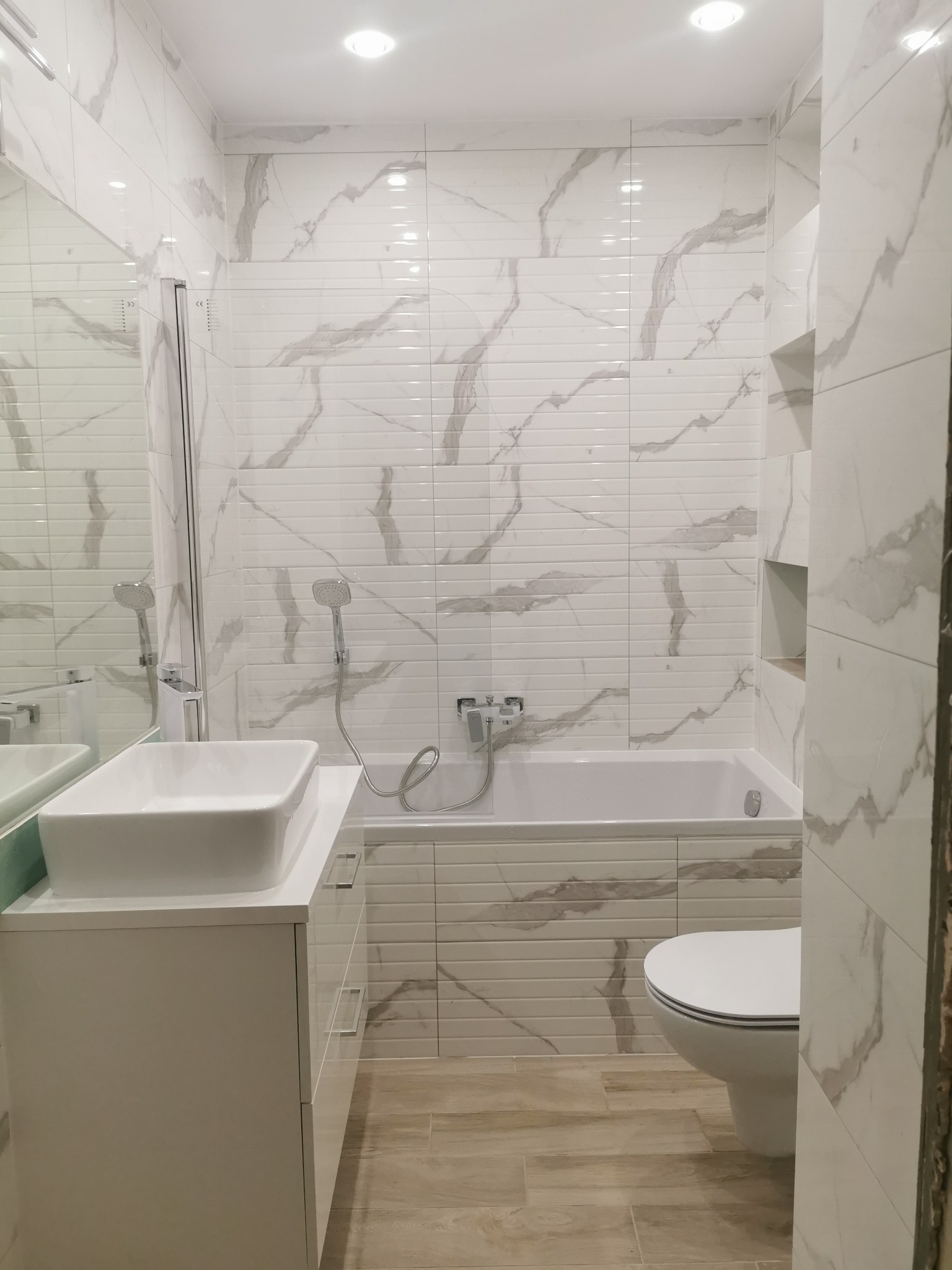 Nowoczesna łazienka z białymi płytkami imitującymi marmur, podłużnymi płytkami nad wanną i drewnopodobną podłogą. Umywalka nablatowa na białej szafce z szufladami, chromowana armatura i lustro.