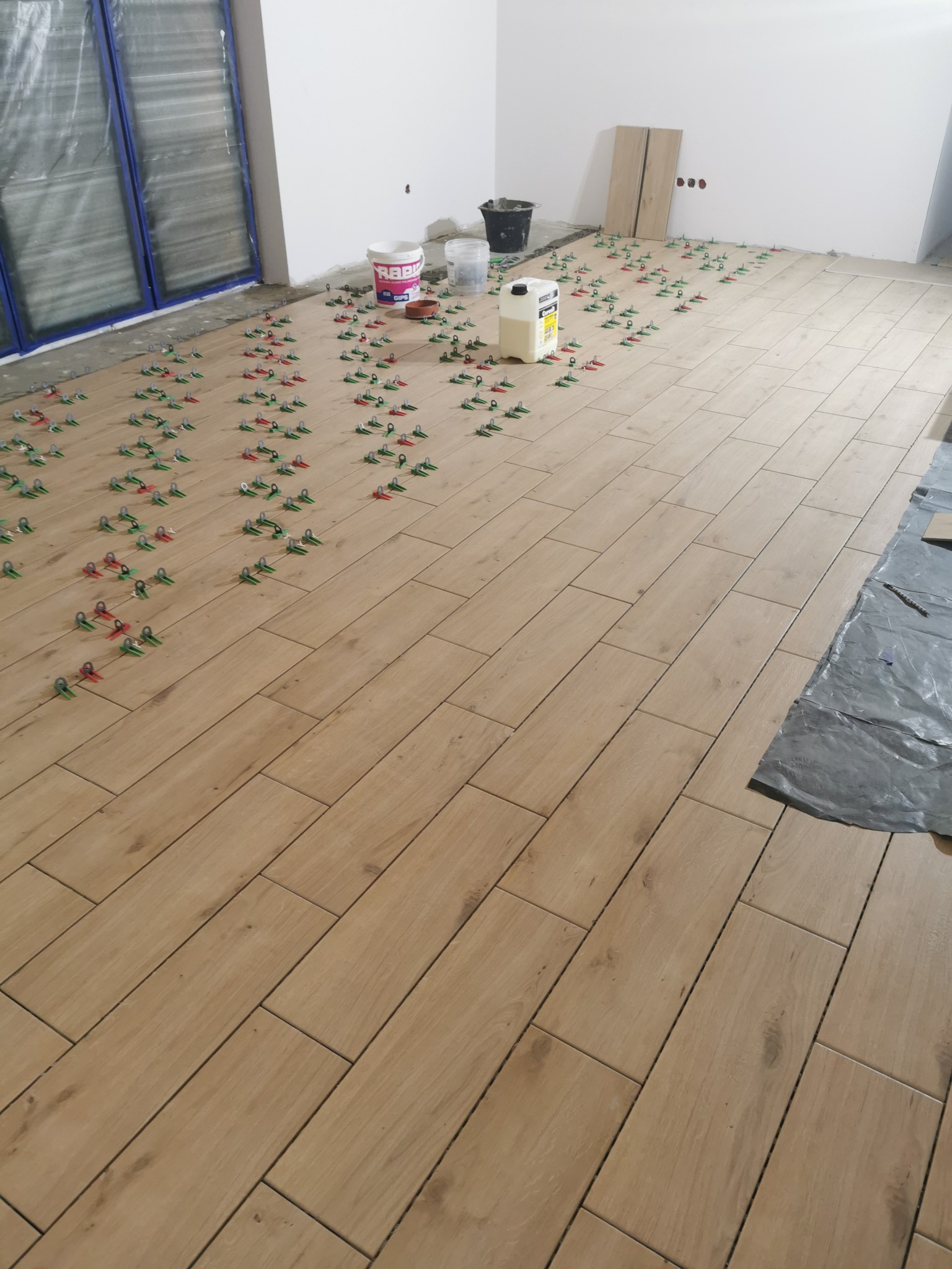 Układanie płytek podłogowych imitujących drewno, z użyciem klipsów poziomujących, widoczne wiaderka i pojemnik z klejem w jasnym, surowym wnętrzu.