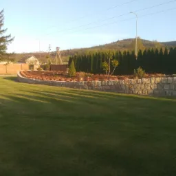 Ogród z niskim murem z jasnej kostki granitowej, za nim nasadzenia krzewów i drzew, w tle budynek i zalesione wzgórze, trawnik z długimi cieniami.