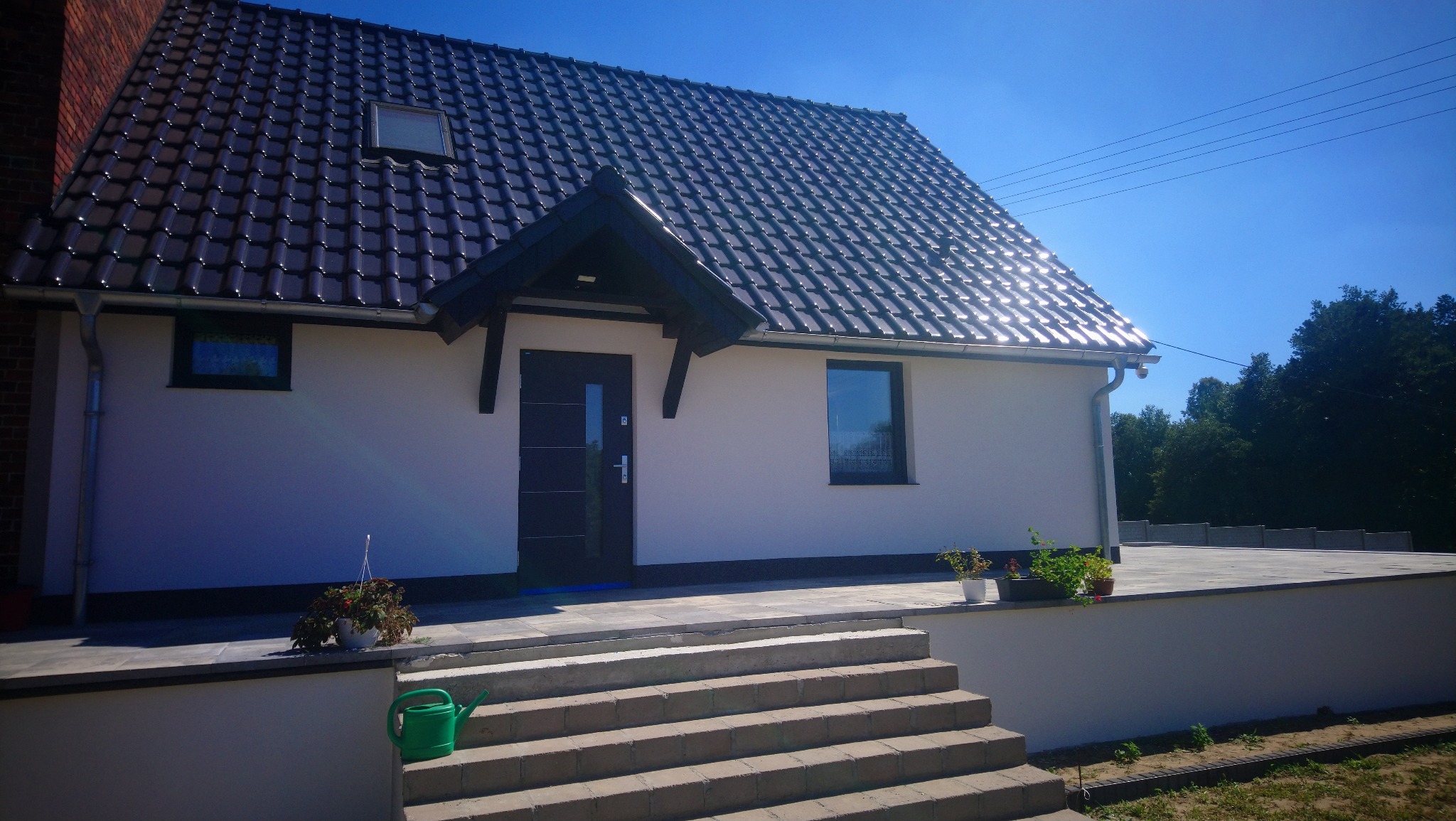 Nowo wybudowany dom jednorodzinny z ciemną dachówką i stolarką okienną, widok z przodu, z zielonym dzbankiem do podlewania na schodach.