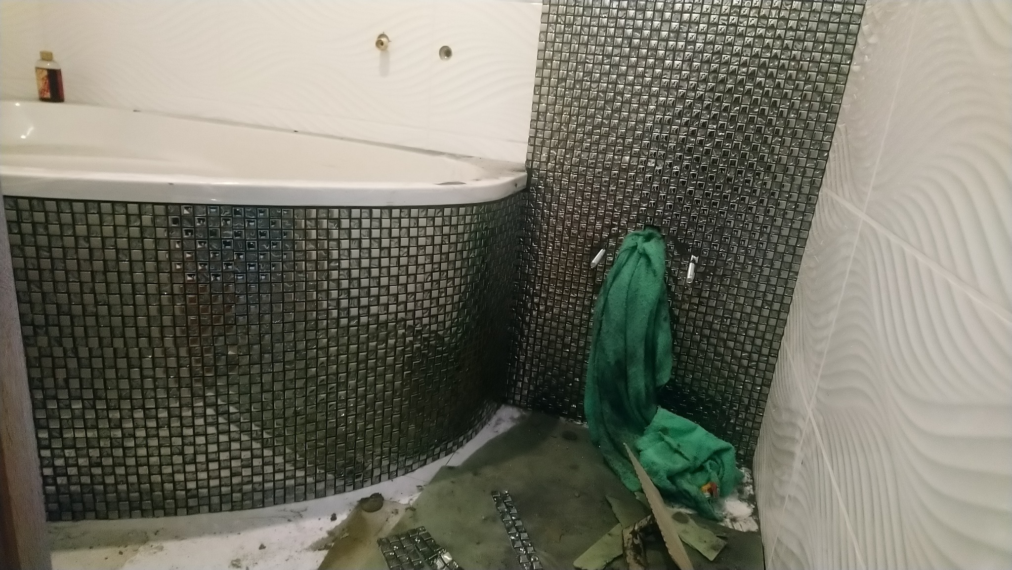 Montaż mozaiki na obudowie wanny narożnej i ścianie łazienki, widoczne niedociągnięcia i resztki kleju na podłodze, zielony ręcznik na haczyku.