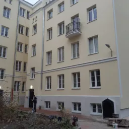 Dziedziniec kamienicy po remoncie elewacji, widoczne świeżo otynkowane i pomalowane ściany w odcieniach beżu, pracownik myjący chodnik z resztek farby, worki z cementem na pierwszym planie.