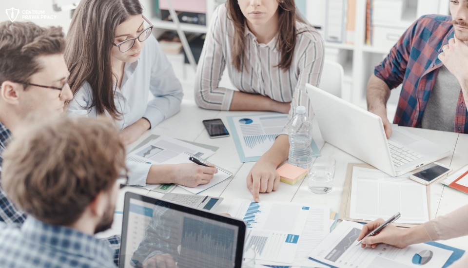 Zespół osób analizuje dokumenty i wykresy na białym stole w jasnym biurze, używając laptopów i notatników.