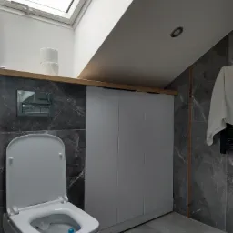 Łazienka na poddaszu z marmurowymi płytkami, białą toaletą z otwartą klapą i szafką pod skosem, oświetlona oknem dachowym. Widoczne niedokończone prace: zwisająca taśma LED na podłodze.