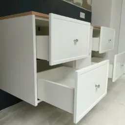 Biała szafka łazienkowa z drewnianym blatem i otwartymi szufladami na tle ciemnej ściany z płytkami i niedokończonej ściany, widoczne gniazdko elektryczne.