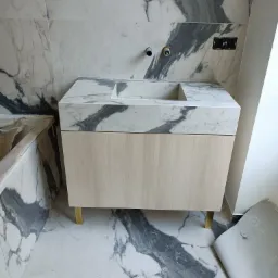 Łazienka w trakcie wykańczania: szafka pod umywalkę z blatem imitującym marmur, drewniane fronty, złote nóżki, marmurowe płytki na ścianach i podłodze, widoczne instalacje i zabezpieczona wanna.