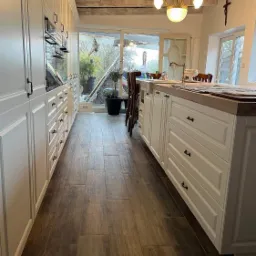 Jasna kuchnia z drewnianą podłogą i blatem, widok na ogród przez duże okno, wisząca lampa w stylu retro, eleganckie fronty szafek z czarnymi uchwytami.