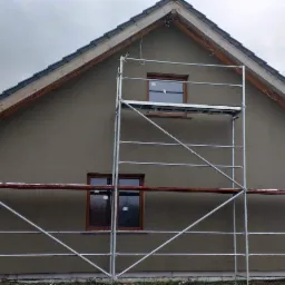 Dom jednorodzinny z szarą elewacją w trakcie prac wykończeniowych, widoczne rusztowanie i fragmenty śniegu na ziemi.