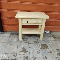 Surowa, jasna szafka nocna z szufladą i półką, stojąca na kostce brukowej przed drewnianą ścianą, widoczne ślady wilgoci na kostce.