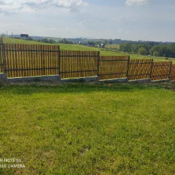 Nowoczesne ogrodzenie panelowe z drewnianymi sztachetami i betonową podmurówką, widok na pagórkowaty krajobraz z domami w tle.