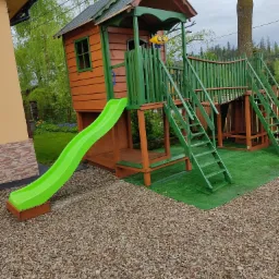 Drewniany plac zabaw z domkiem, zjeżdżalnią i schodami, pomalowany na brązowo i zielono, umieszczony na sztucznej trawie i żwirowym podłożu.