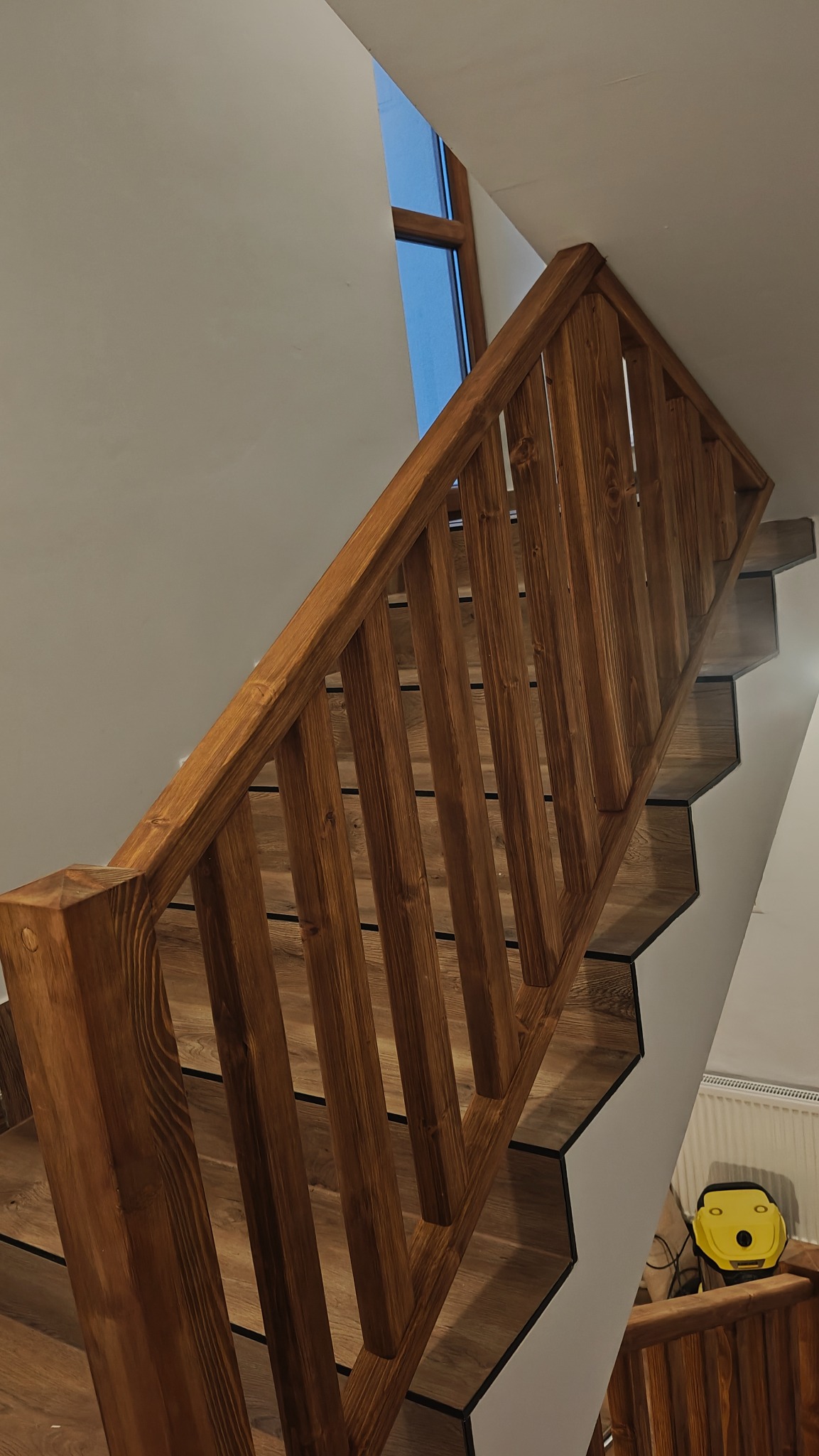 Klatka schodowa z drewnianą balustradą w ciepłym odcieniu brązu. Widoczne stopnie wykończone panelami imitującymi drewno. Ujęcie z dołu, w tle okno.