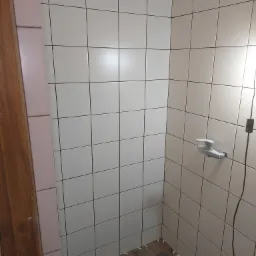 Ściana w trakcie układania jasnych, kwadratowych płytek ceramicznych w łazience, widoczne krzyżyki dystansowe na podłodze i fragment instalacji wodnej.