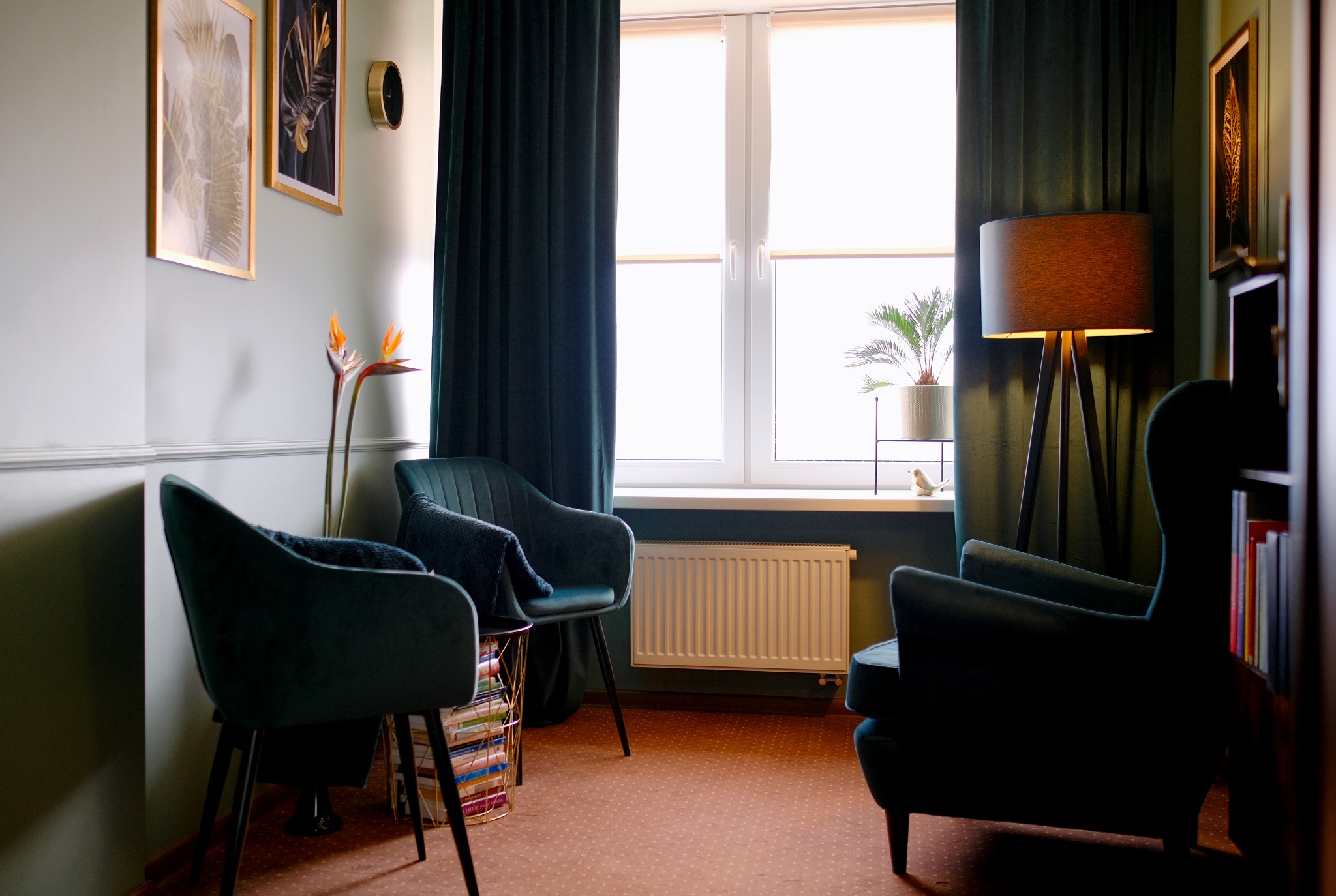 Wnętrze gabinetu z dwoma welurowymi fotelami w odcieniach zieleni, lampą stojącą na trójnogu, obrazami na ścianie i kwiatem strelicji królewskiej w wazonie.