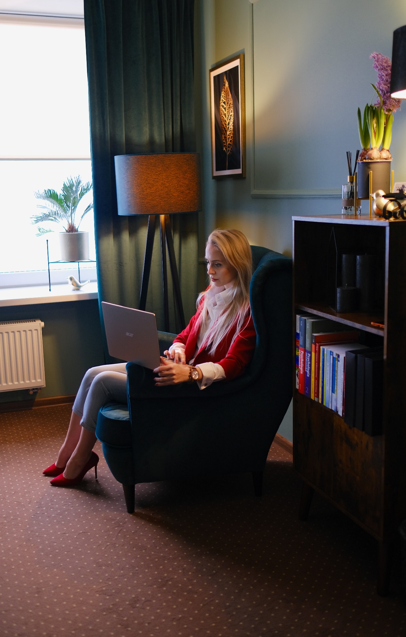 Kobieta w czerwonej marynarce siedzi w fotelu i pracuje na laptopie w gabinecie z biblioteczką.