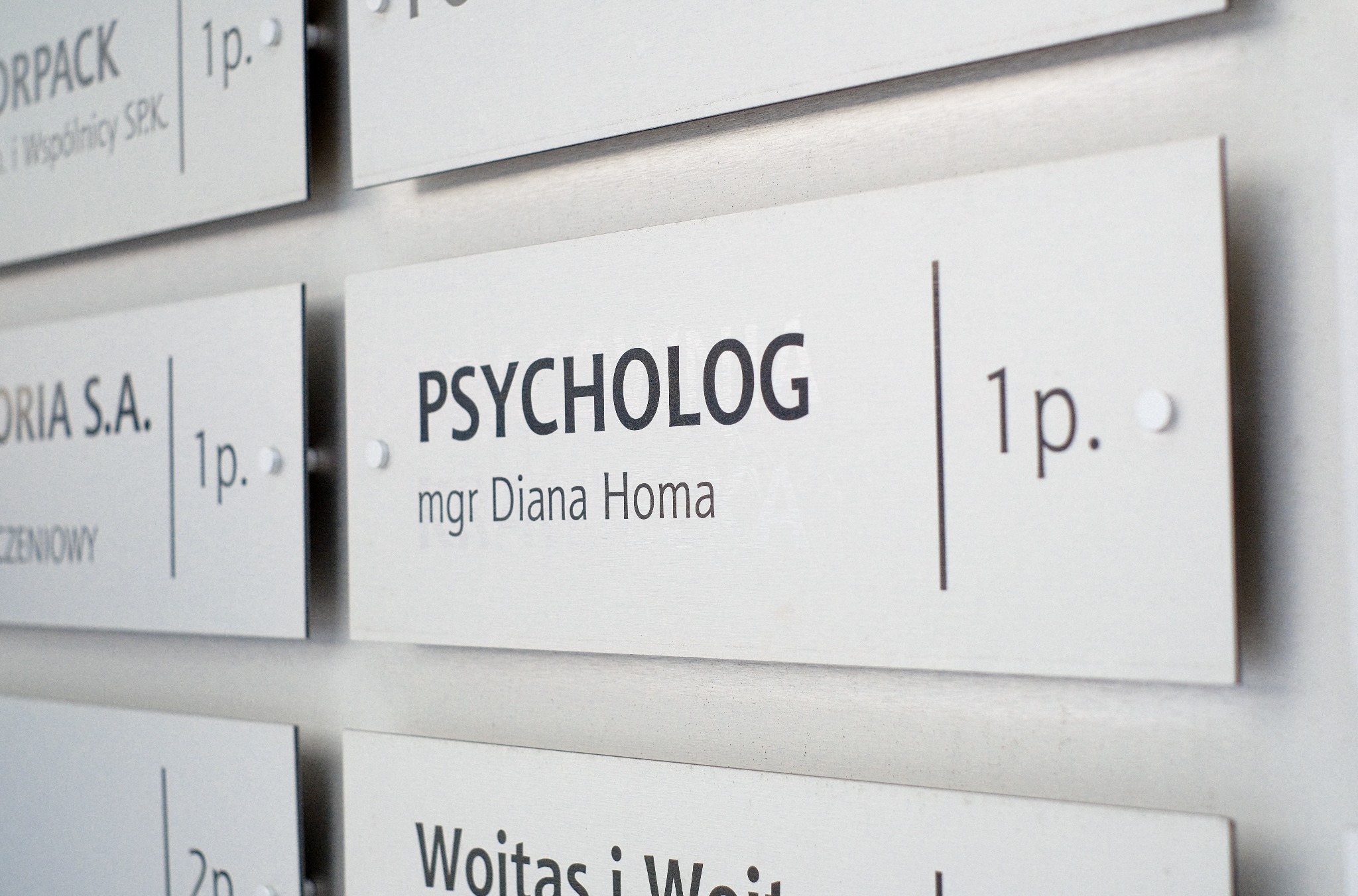 Tabliczka informacyjna na ścianie z napisem 'PSYCHOLOG mgr Diana Homa' umieszczona obok innych tabliczek z nazwami firm i numerami pięter.