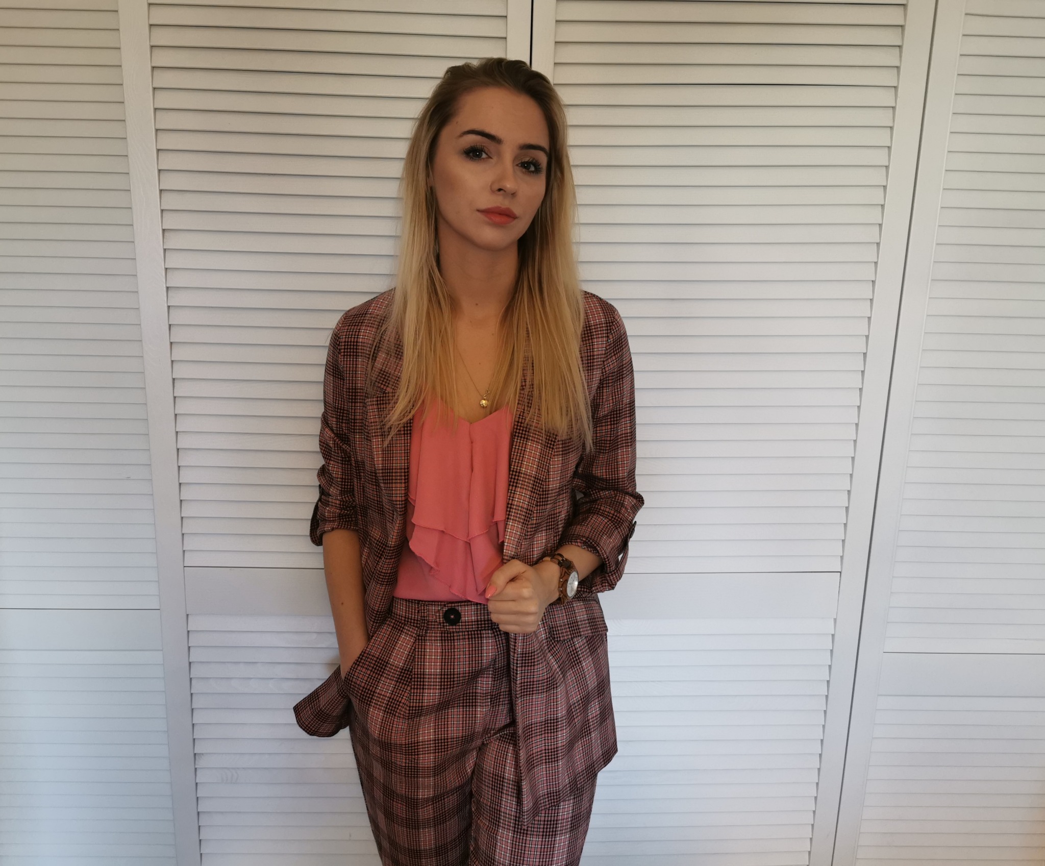 Portret kobiety w różowym garniturze w kratę na tle białych żaluzji.