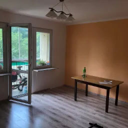 Pokój przed odświeżeniem: podłoga z paneli, pomarańczowa ściana, stół z butelką wody, otwarte drzwi balkonowe z widokiem na zieleń.