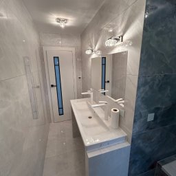 PROFBUD Wykończenia Wnętrz Patryk Lasota - Nowoczesna łazienka z dwiema umywalkami nablatowymi, lustrem z kryształowymi lampami, szarą marmurową okładziną ścienną oraz drzwiami z niebieskim akcentem.