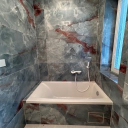 PROFBUD Wykończenia Wnętrz Patryk Lasota - Wykończona łazienka z wanną obłożoną płytkami imitującymi marmur w odcieniach szarości i czerwieni, widoczna armatura i okno z żaluzjami.