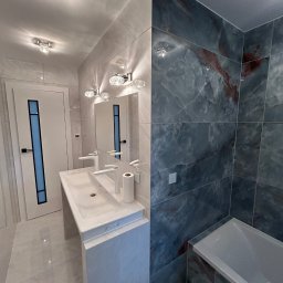 PROFBUD Wykończenia Wnętrz Patryk Lasota - Nowoczesna łazienka z białą umywalką nablatową, baterią w kształcie litery C, lustrem z oświetleniem, drzwiami z niebieskim akcentem i płytkami imitującymi marmur w odcieniach szarości i błękitu.