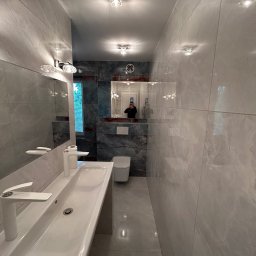 PROFBUD Wykończenia Wnętrz Patryk Lasota - Wąska, wykończona łazienka z szarymi płytkami imitującymi marmur na ścianach i podłodze, biała umywalka nablatowa z nowoczesną baterią, lustro z oświetleniem i odbiciem osoby robiącej zdjęcie...