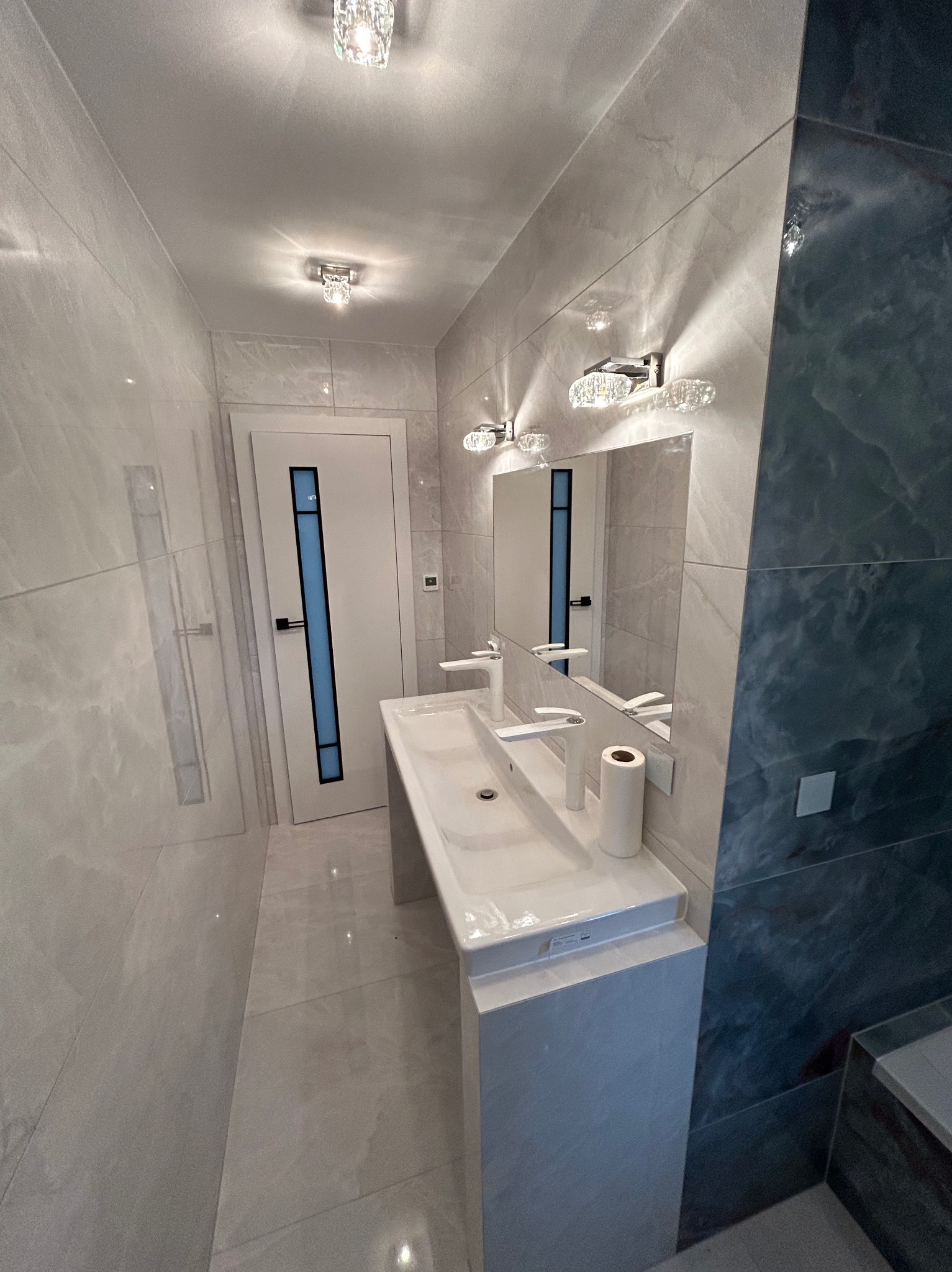 Nowoczesna łazienka z dwiema umywalkami nablatowymi, lustrem z kryształowymi lampami, szarą marmurową okładziną ścienną oraz drzwiami z niebieskim akcentem.