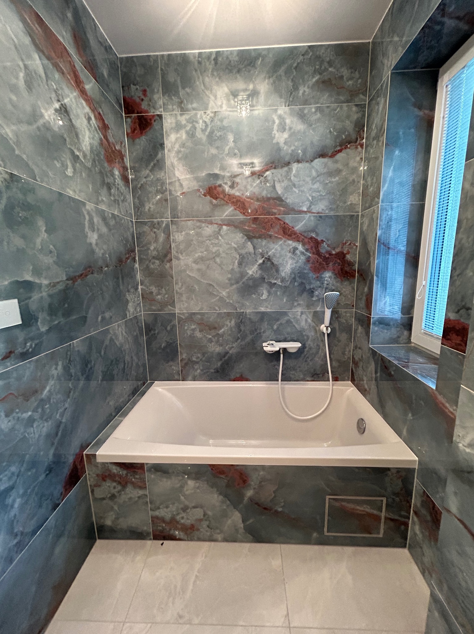 Wykończona łazienka z wanną obłożoną płytkami imitującymi marmur w odcieniach szarości i czerwieni, widoczna armatura i okno z żaluzjami.