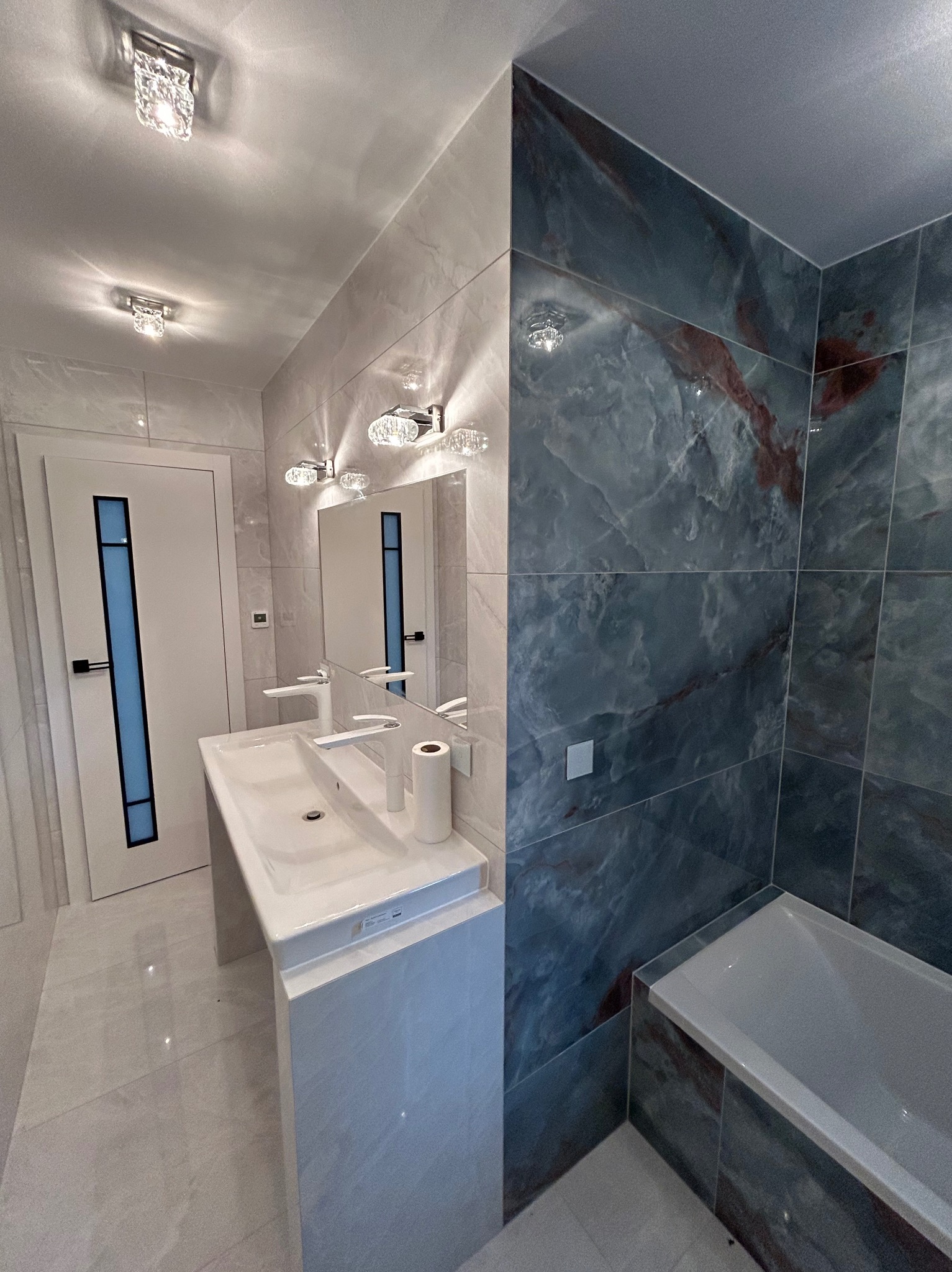 Nowoczesna łazienka z białą umywalką nablatową, baterią w kształcie litery C, lustrem z oświetleniem, drzwiami z niebieskim akcentem i płytkami imitującymi marmur w odcieniach szarości i błękitu.